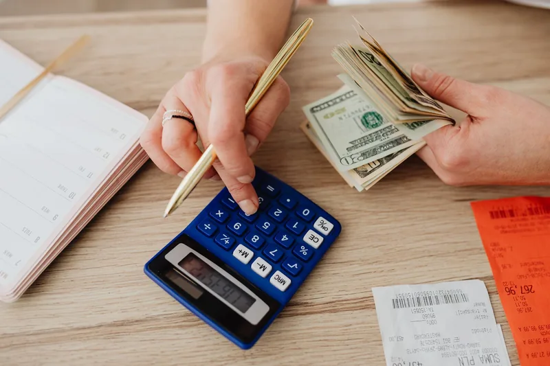Person adding up costs on a calculator with receipts, cash and a notebook on a desk