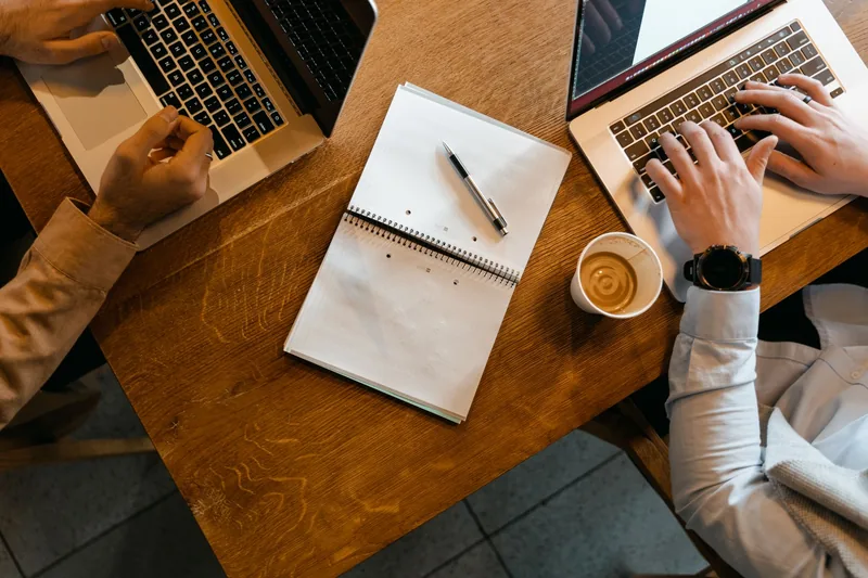 Two people working side by side on laptops at a wooden table, reviewing an ad account together